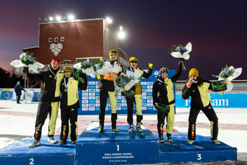 Sechs Personen auf einem Podium. Sie tragen Wintersport-Kleidung, haben Medaillen um den Hals hängen und halten Blumensträuße in den Händen. Auf der Vorderseite des Podiums stehen die Zahlen eins, zwei und drei. In der Mitte steht außerdem "Para Nordic Skiing Wolrd Championships Östersund".