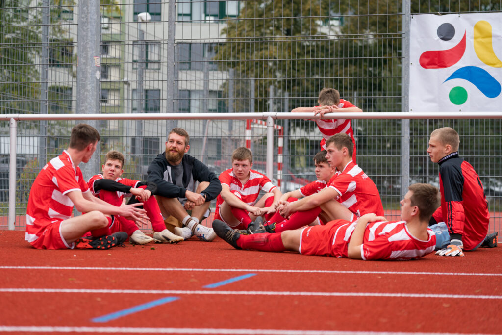 Eine Gruppe von Menschen in roten Trikots sitzt auf einer Tartanbahn. in der rechten oberen Ecke des Bildes ist das Logo der SportWoche für Alle zu sehen.